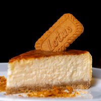 Slice of creamy cheesecake with a Lotus Biscoff biscuit topper on a white plate.